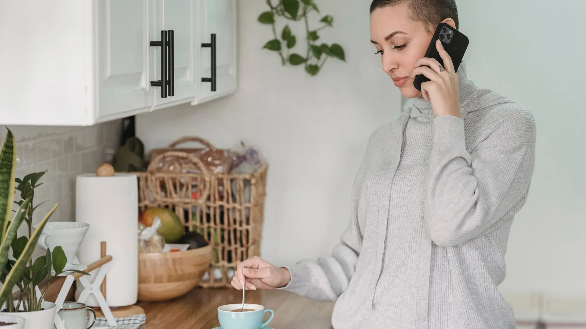Woman in a grey jumper on a phone call while stirring coffee in a bright kitchen